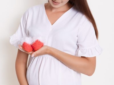 Gently pregnant woman with red booties, touching her belly, expectant mother with baby's booties posing against white wall, brunette lady with long beautiful expecting child.